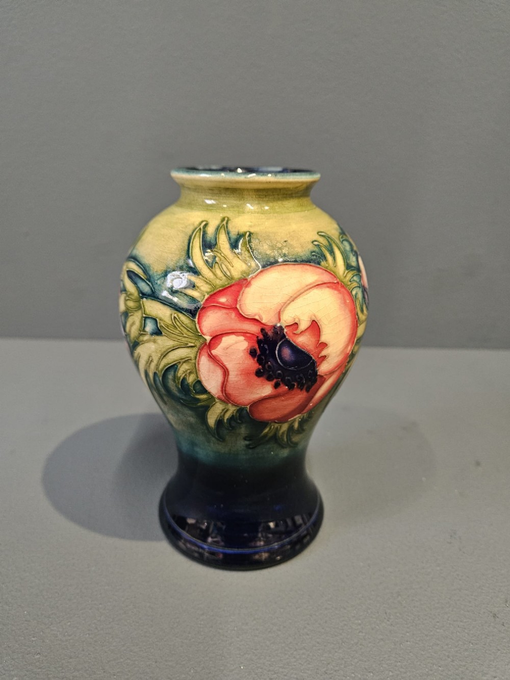 A small baluster-shaped ceramic vase featuring a tubelined red anemone motif against a ground fading from yellow-green to dark blue.