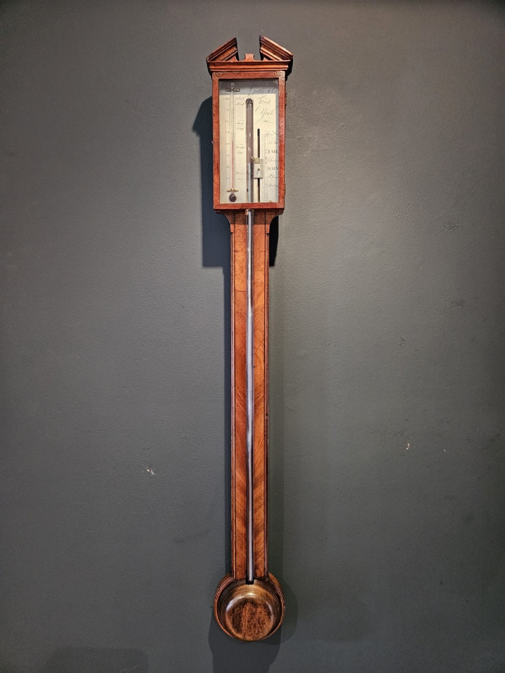 A wall-mounted Georgian mahogany stick barometer featuring a broken-arch pediment, a silvered register plate behind glass, and a turned cistern cover at the base.