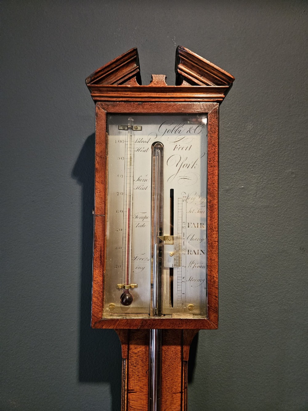 A wall-mounted Georgian mahogany stick barometer featuring a broken-arch pediment, a silvered register plate behind glass, and a turned cistern cover at the base. View III