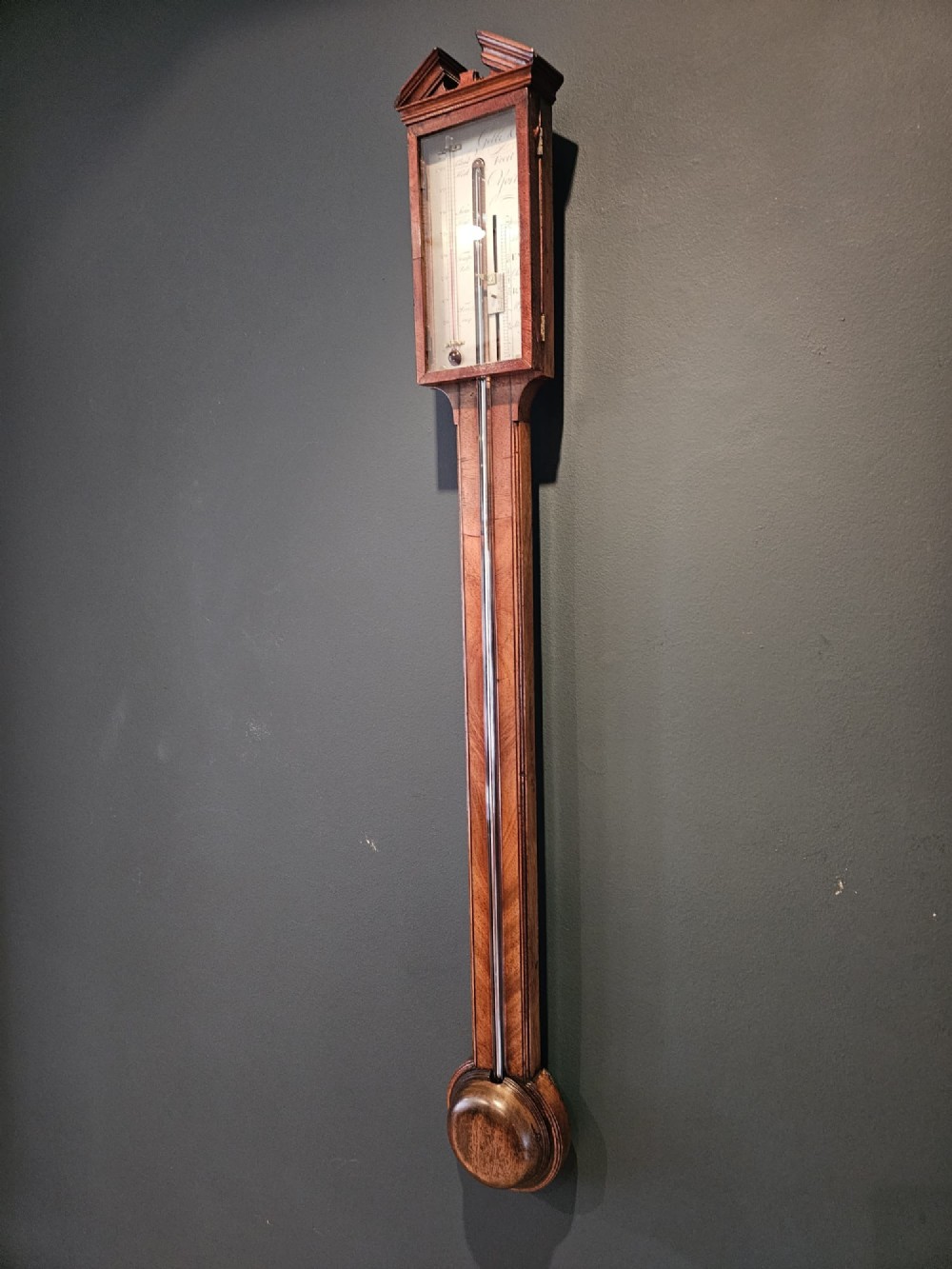 A wall-mounted Georgian mahogany stick barometer featuring a broken-arch pediment, a silvered register plate behind glass, and a turned cistern cover at the base. View IV
