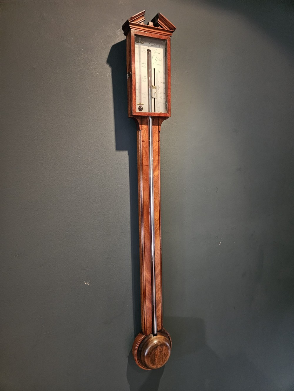 A wall-mounted Georgian mahogany stick barometer featuring a broken-arch pediment, a silvered register plate behind glass, and a turned cistern cover at the base. View VI