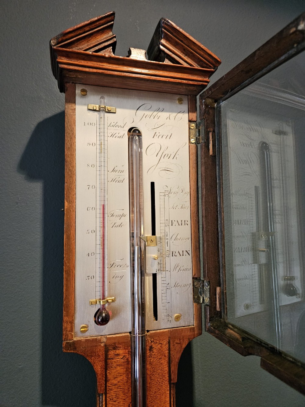 A wall-mounted Georgian mahogany stick barometer featuring a broken-arch pediment, a silvered register plate behind glass, and a turned cistern cover at the base. View VII