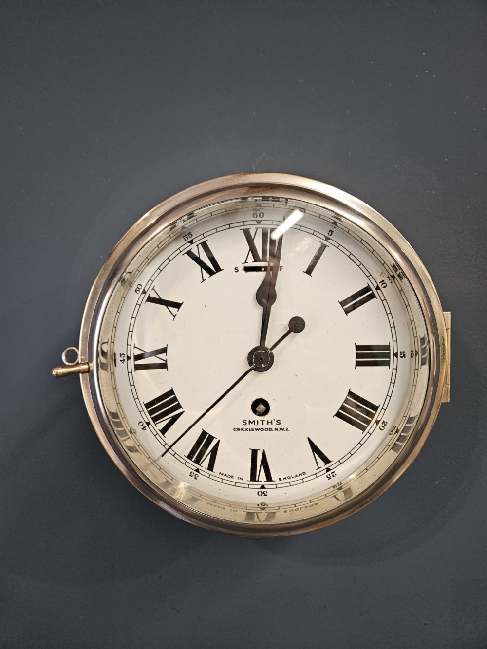 An antique circular brass ships clock with a white enamel dial, black Roman numerals, and a central seconds sweep hand. View V