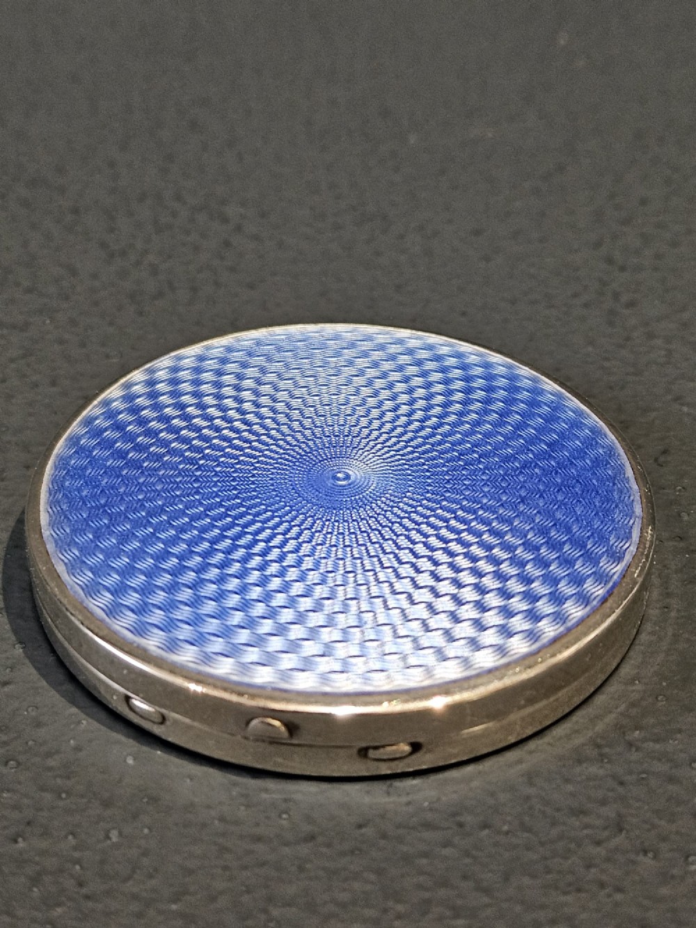 A circular silver compact featuring a vibrant blue engine-turned guilloche enamel lid with a radiating pattern.