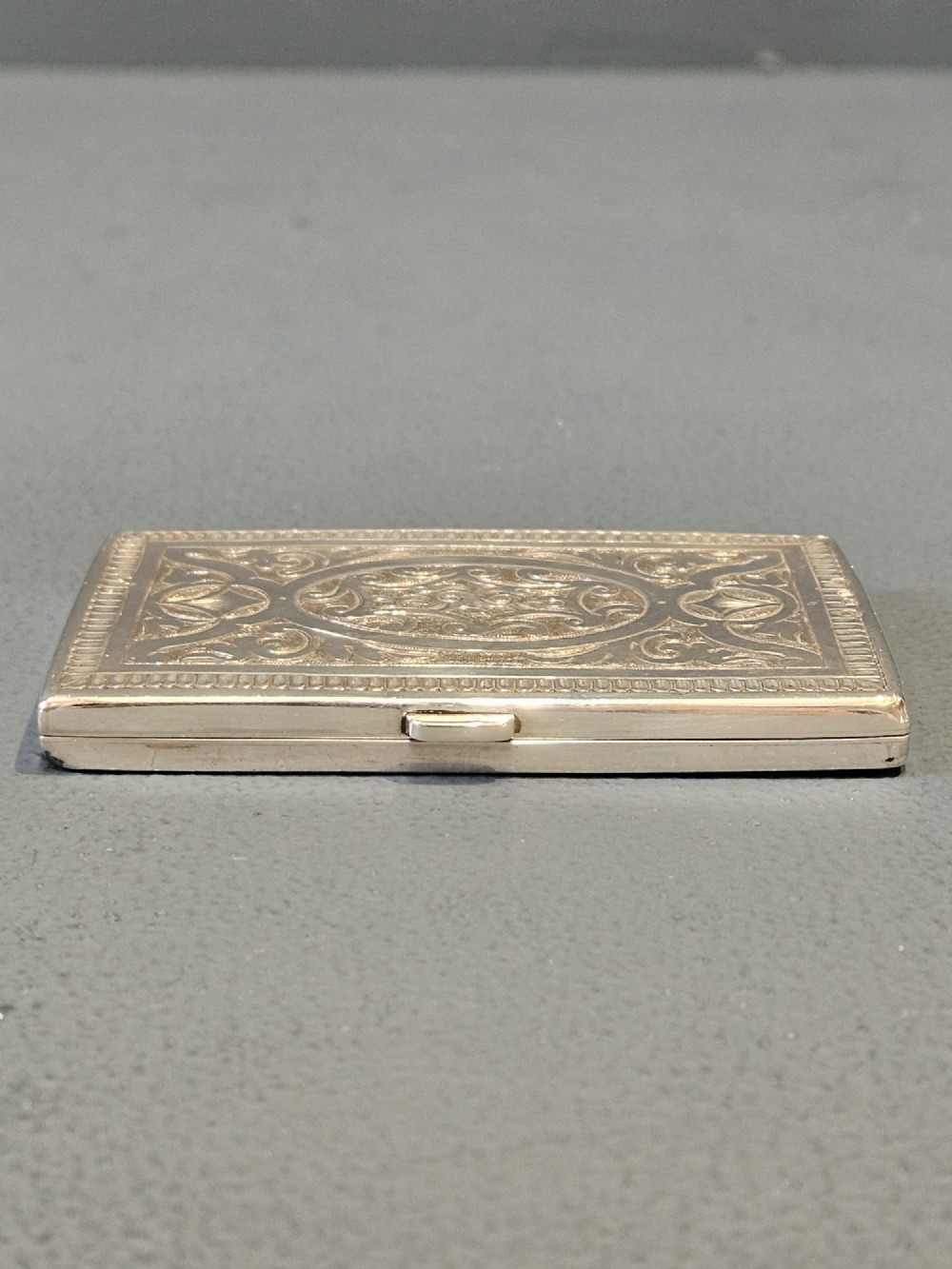 A rectangular Victorian silver card case featuring hand-engraved scrolling foliage, an oval central medallion, and a hinged side edge. View V