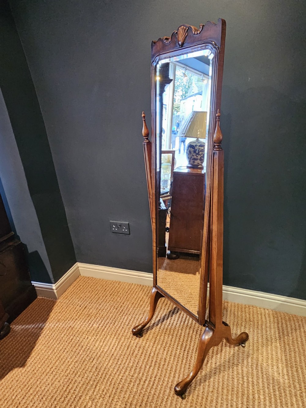 A George III style mahogany cheval mirror featuring an arched top with a carved shell cresting, turned side finials, and splayed legs on brass castors. View VI