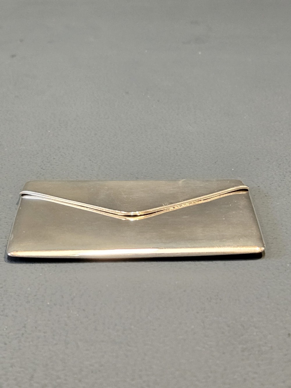 An antique sterling silver envelope-shaped card case with visible hallmarks along the flap seam resting against a dark grey backdrop. View II