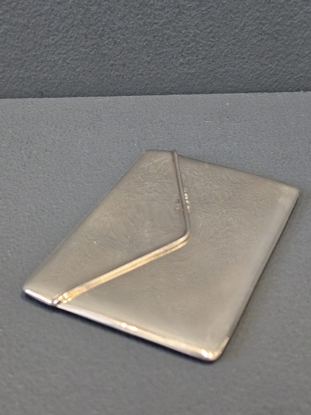 An antique sterling silver envelope-shaped card case with visible hallmarks along the flap seam resting against a dark grey backdrop. View IV