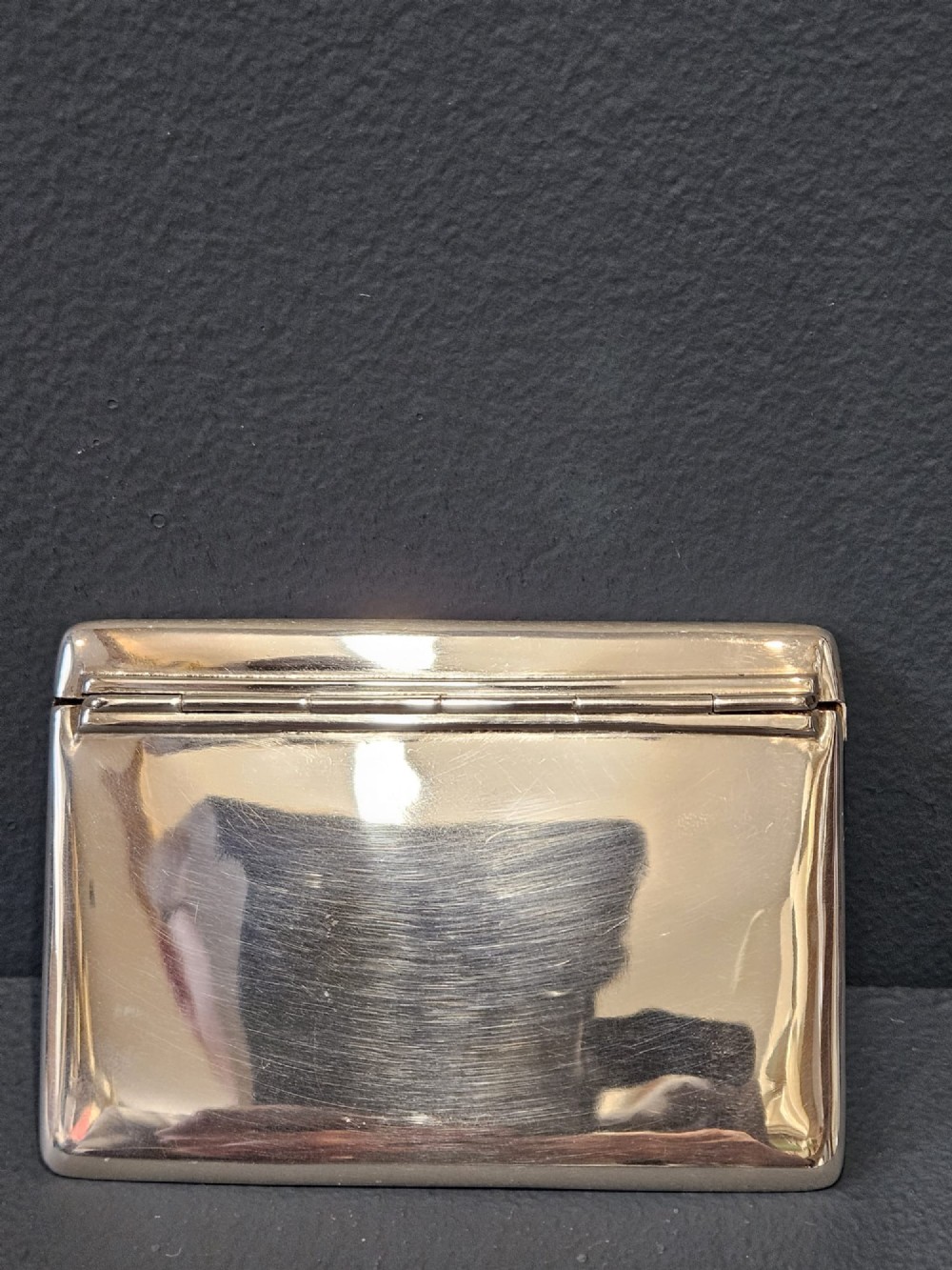An antique sterling silver envelope-shaped card case with visible hallmarks along the flap seam resting against a dark grey backdrop. View V