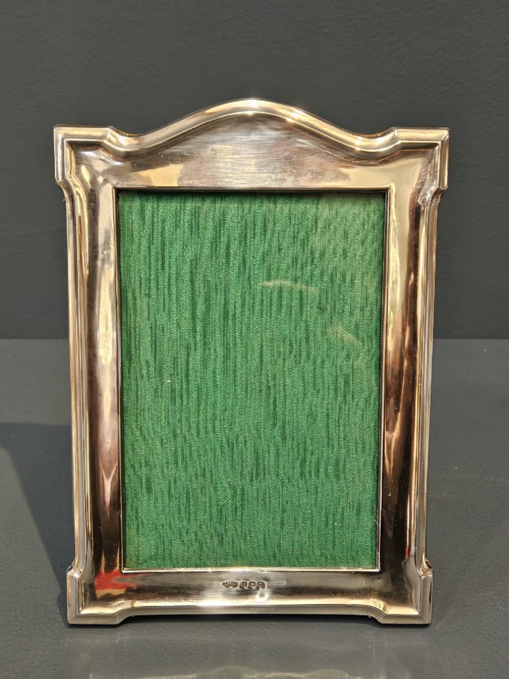 A sterling silver photo frame featuring a swept arched top, moulded border, visible hallmarks at the base, and a green fabric backing.