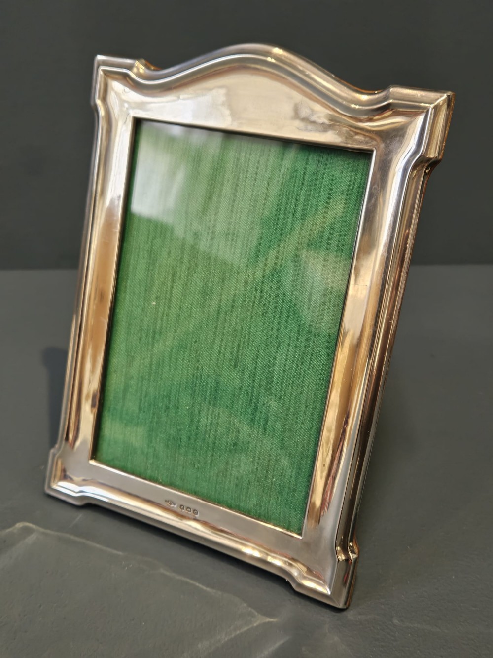 A sterling silver photo frame featuring a swept arched top, moulded border, visible hallmarks at the base, and a green fabric backing. View II