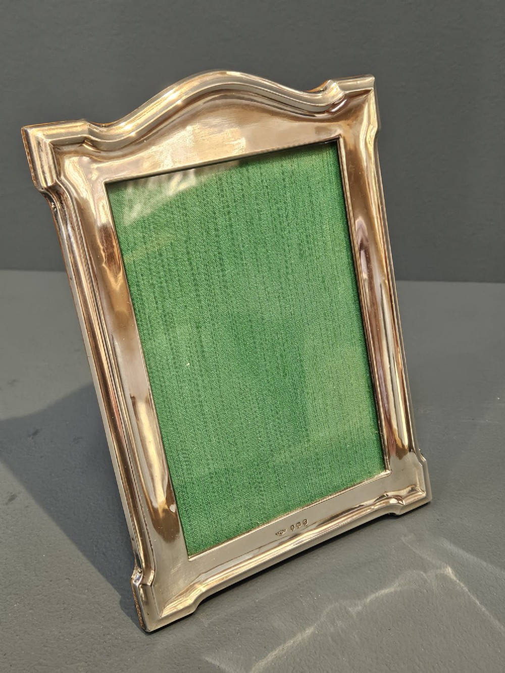 A sterling silver photo frame featuring a swept arched top, moulded border, visible hallmarks at the base, and a green fabric backing. View IV