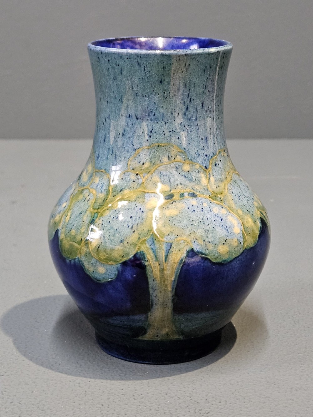A small baluster-shaped ceramic vase featuring tubelined tree motifs in tones of green and turquoise over a deep cobalt blue glaze. View II