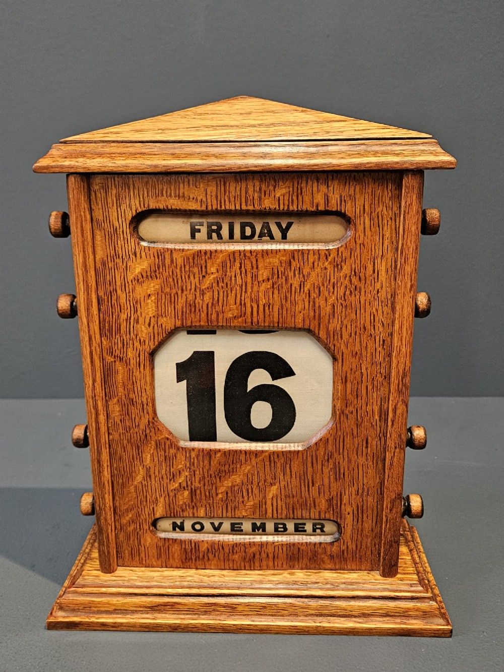 An Edwardian figured oak perpetual desk calendar featuring a pediment top, moulded base, three display apertures, and turned wooden side knobs. View III