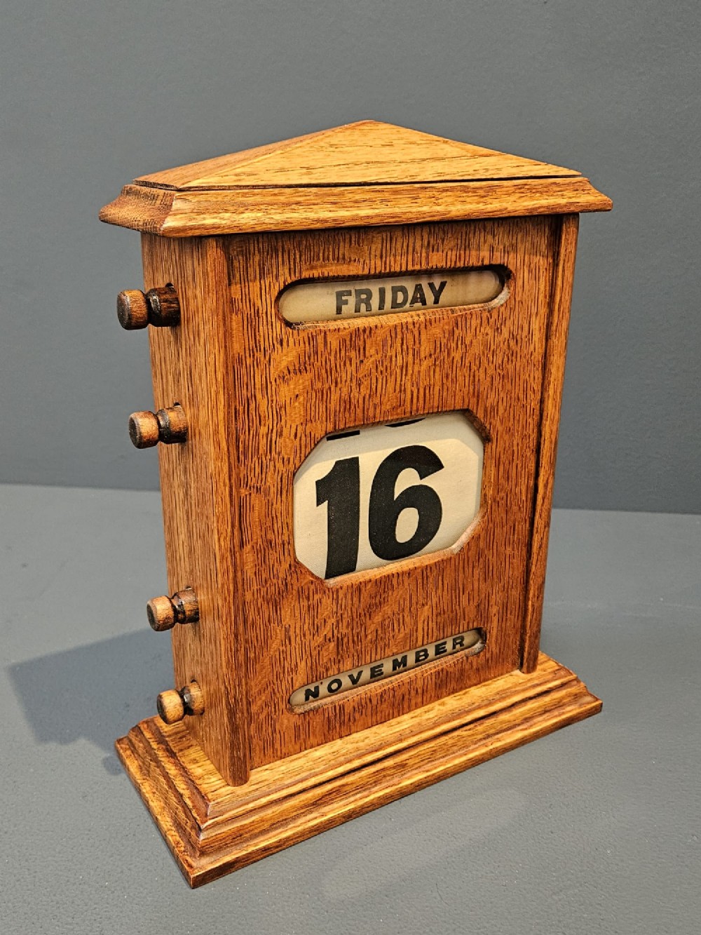 An Edwardian figured oak perpetual desk calendar featuring a pediment top, moulded base, three display apertures, and turned wooden side knobs. View IV