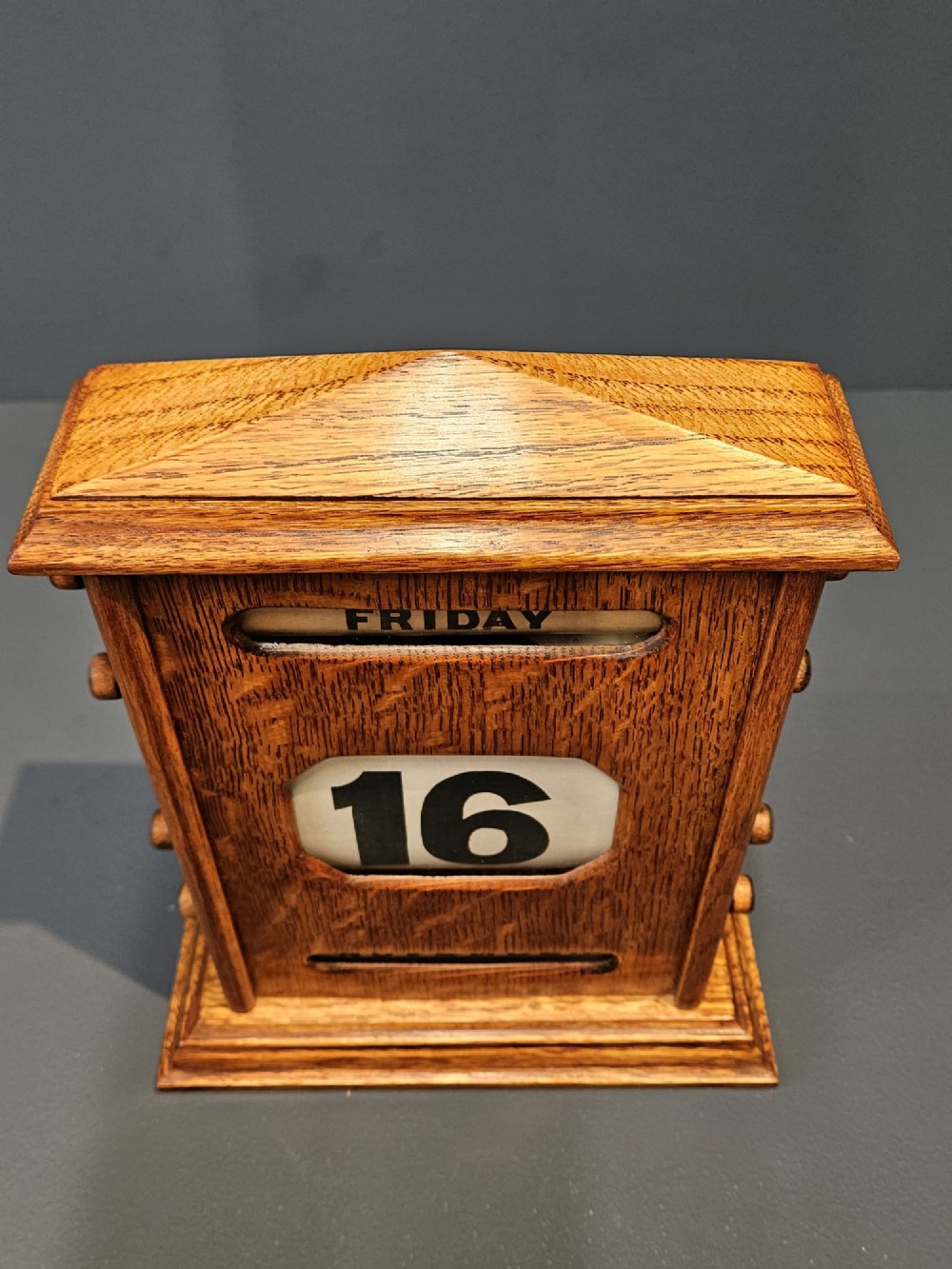 An Edwardian figured oak perpetual desk calendar featuring a pediment top, moulded base, three display apertures, and turned wooden side knobs. View V