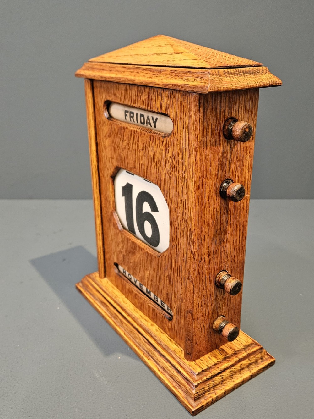 An Edwardian figured oak perpetual desk calendar featuring a pediment top, moulded base, three display apertures, and turned wooden side knobs. View VI