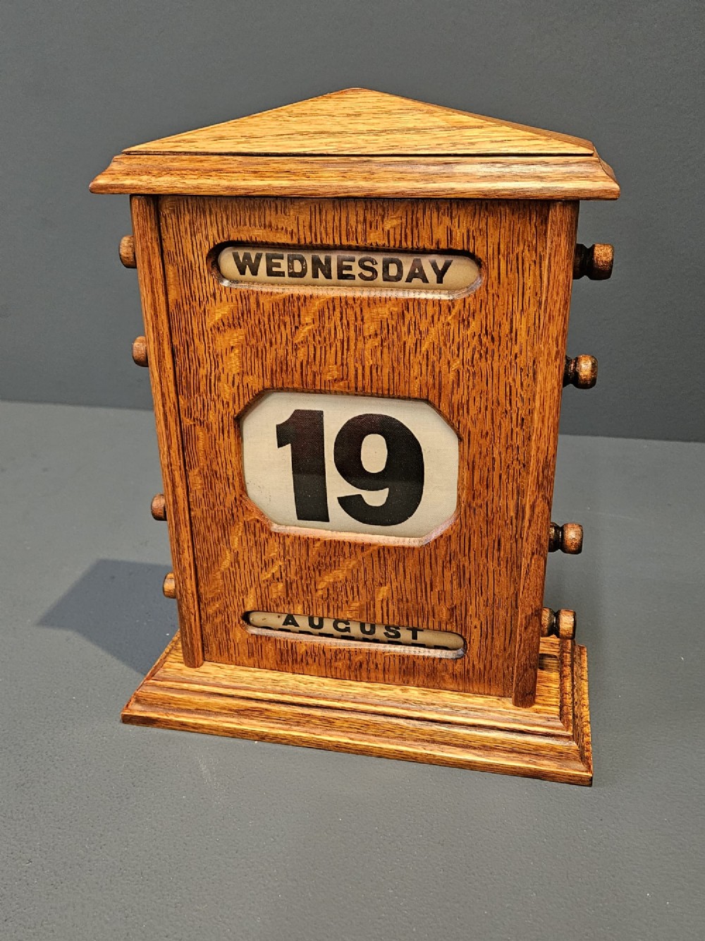 An Edwardian figured oak perpetual desk calendar featuring a pediment top, moulded base, three display apertures, and turned wooden side knobs. View VIII