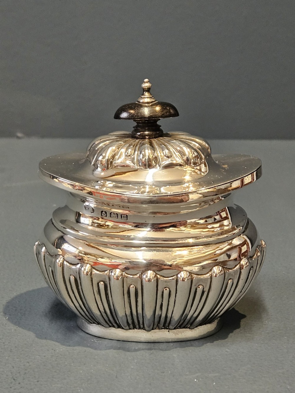 A George V polished sterling silver tea caddy featuring a fluted lower body, hinged lid, ebonised finial, and stamped hallmarks.