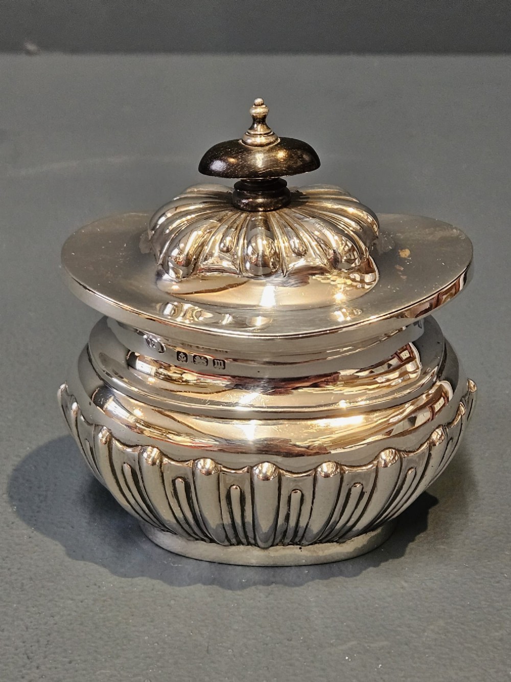 A George V polished sterling silver tea caddy featuring a fluted lower body, hinged lid, ebonised finial, and stamped hallmarks. View II