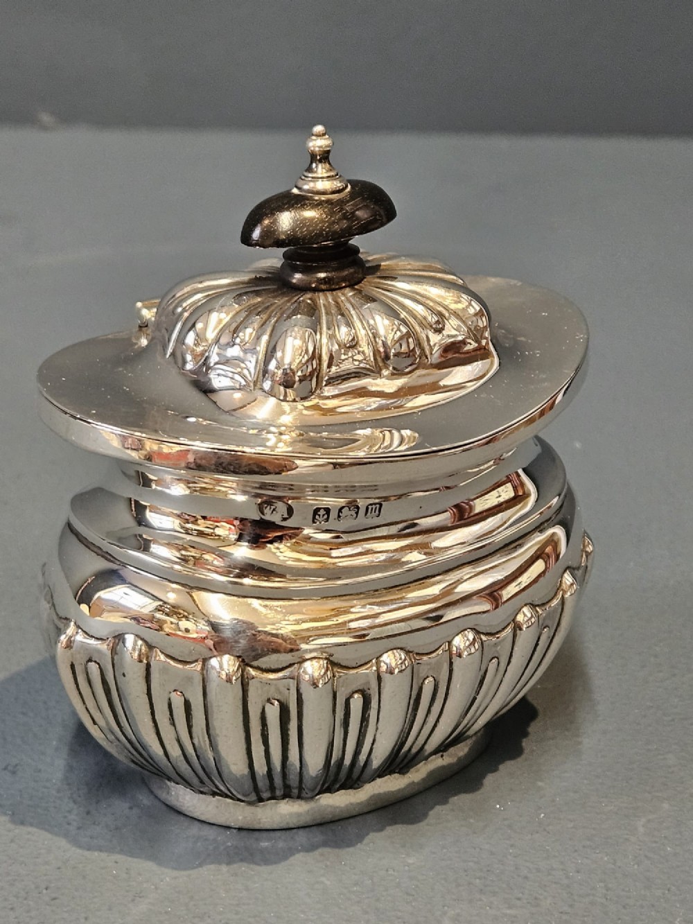 A George V polished sterling silver tea caddy featuring a fluted lower body, hinged lid, ebonised finial, and stamped hallmarks. View III