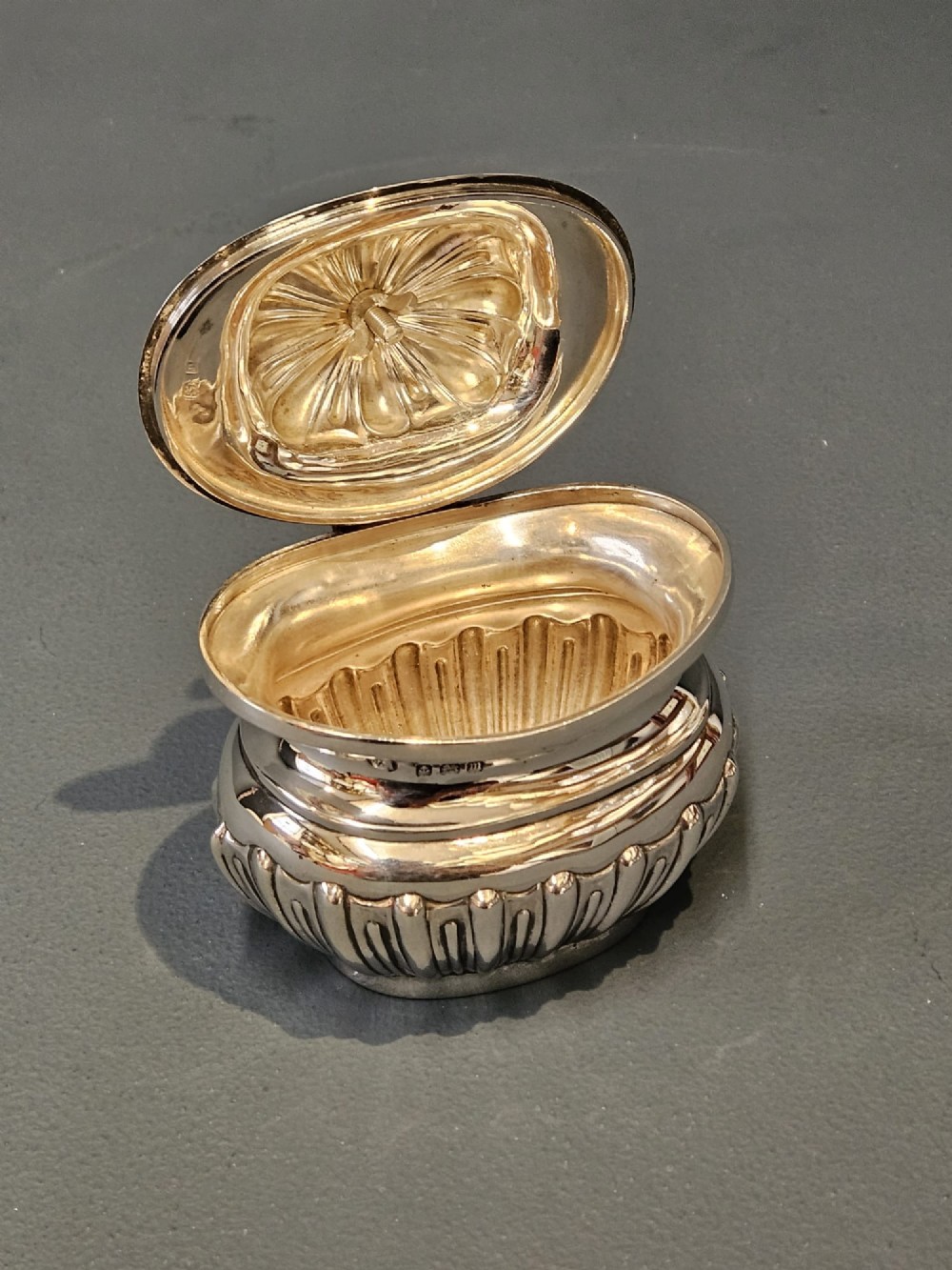 A George V polished sterling silver tea caddy featuring a fluted lower body, hinged lid, ebonised finial, and stamped hallmarks. View VI