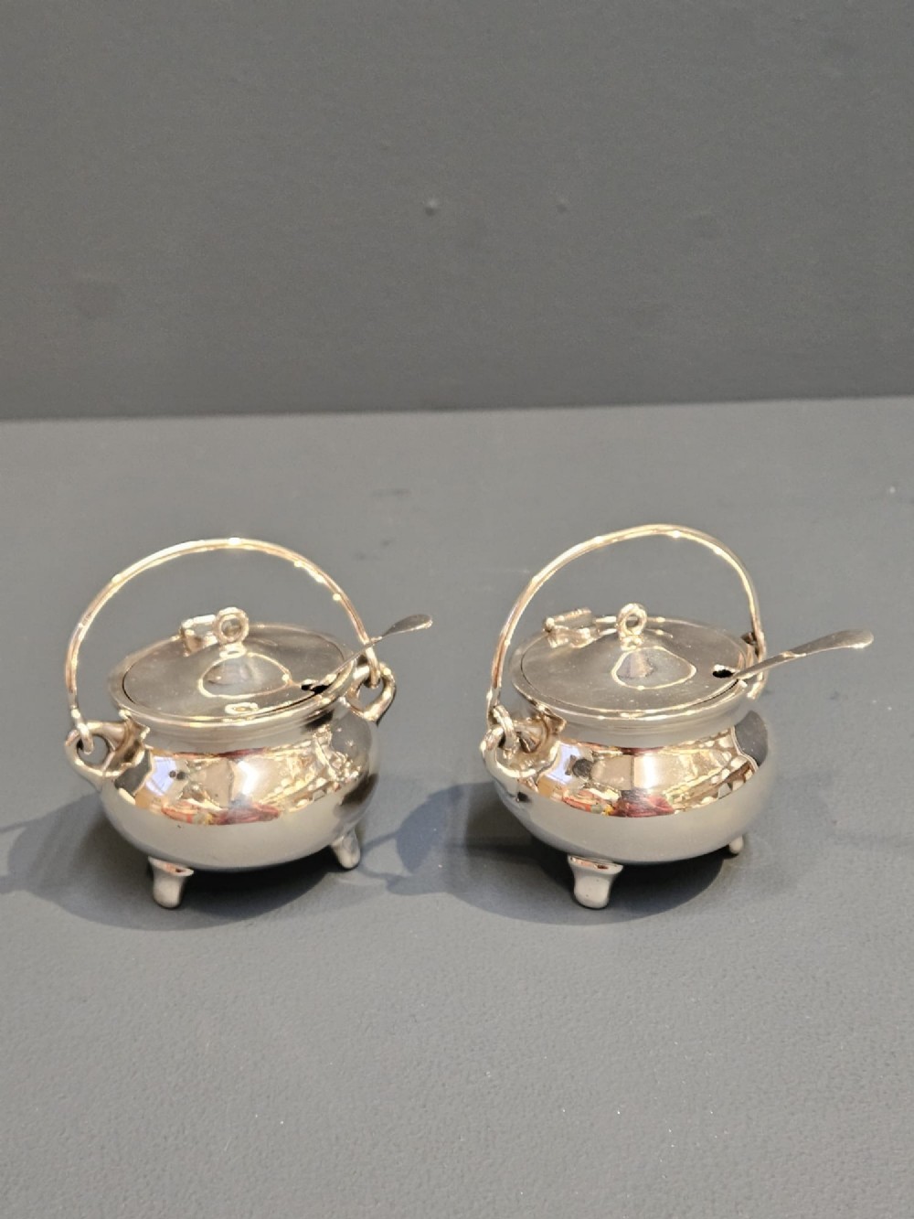 A pair of silver cauldron-shaped Edwardian mustard pots featuring overhead wire handles, hinged lids, and three small feet.