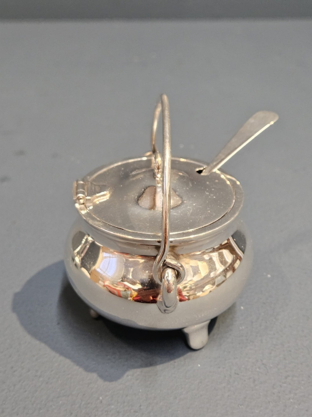 A pair of silver cauldron-shaped Edwardian mustard pots featuring overhead wire handles, hinged lids, and three small feet. View II