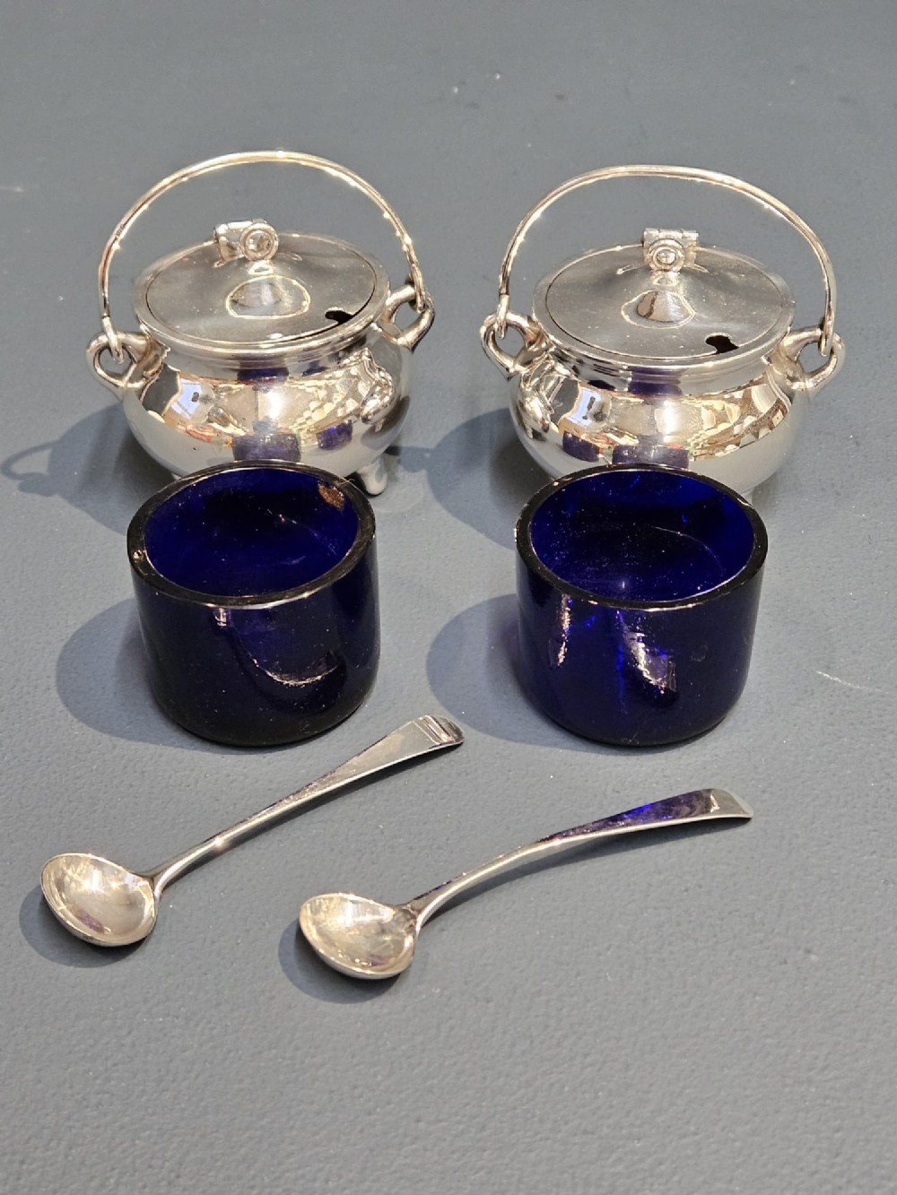 A pair of silver cauldron-shaped Edwardian mustard pots featuring overhead wire handles, hinged lids, and three small feet. View V