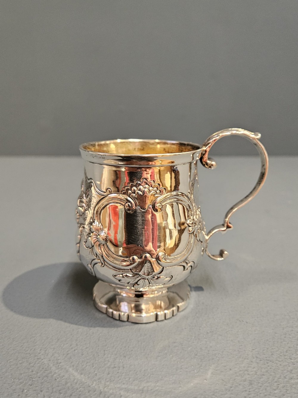 A William IV sterling silver mug featuring hand-chased floral cartouches, a scroll handle, a fluted foot, and a gilded interior.