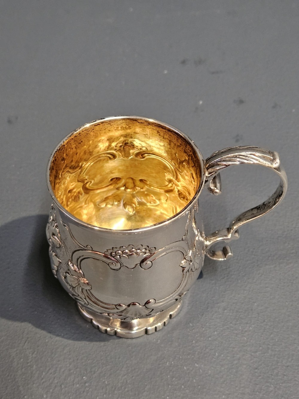 A William IV sterling silver mug featuring hand-chased floral cartouches, a scroll handle, a fluted foot, and a gilded interior. View VI