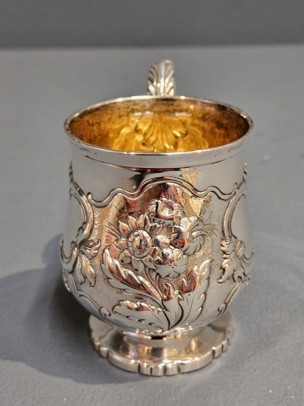 A William IV sterling silver mug featuring hand-chased floral cartouches, a scroll handle, a fluted foot, and a gilded interior. View VIII