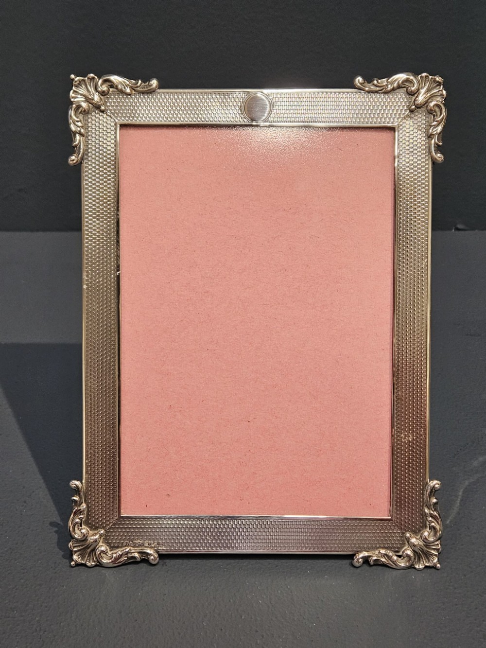 A rectangular George V sterling silver picture frame featuring engine-turned geometric engraving, ornate foliage corner mounts, and a circular top cartouche.
