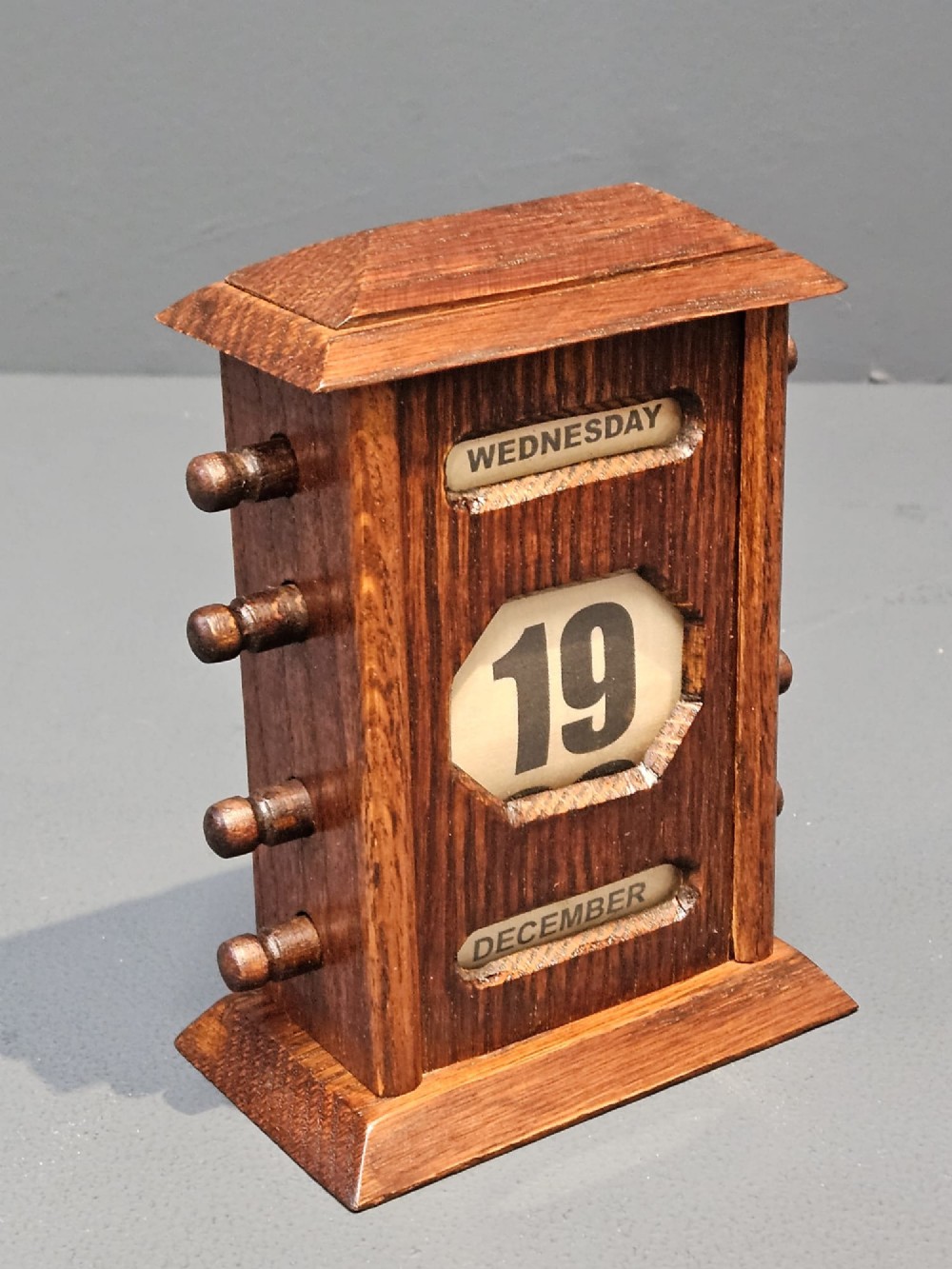 An Edwardian oak perpetual desk calendar featuring turned side knobs and central cutout apertures displaying the day, date, and month on printed fabric scrolls. View II