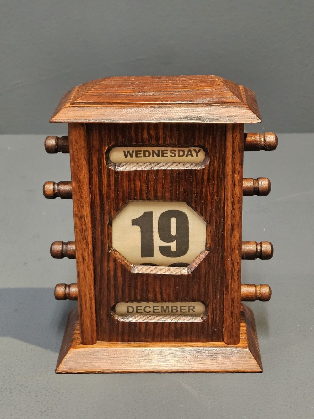 An Edwardian oak perpetual desk calendar featuring turned side knobs and central cutout apertures displaying the day, date, and month on printed fabric scrolls. View V