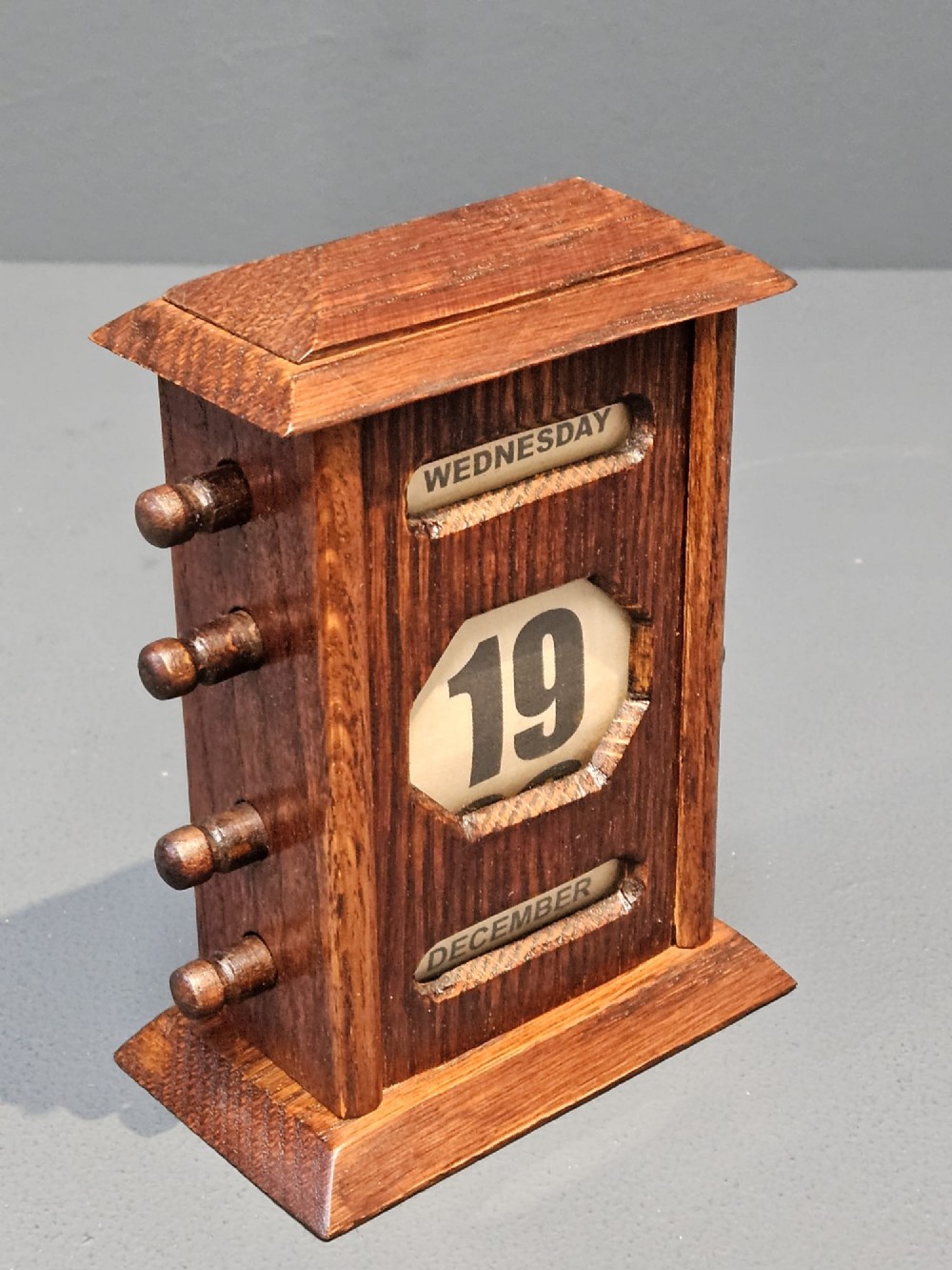 An Edwardian oak perpetual desk calendar featuring turned side knobs and central cutout apertures displaying the day, date, and month on printed fabric scrolls. View VI