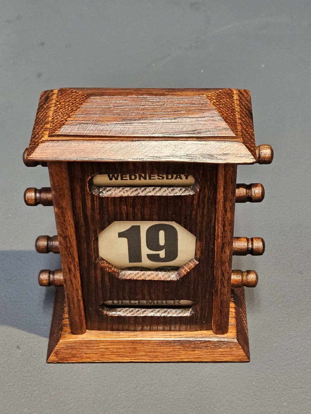 An Edwardian oak perpetual desk calendar featuring turned side knobs and central cutout apertures displaying the day, date, and month on printed fabric scrolls. View VII