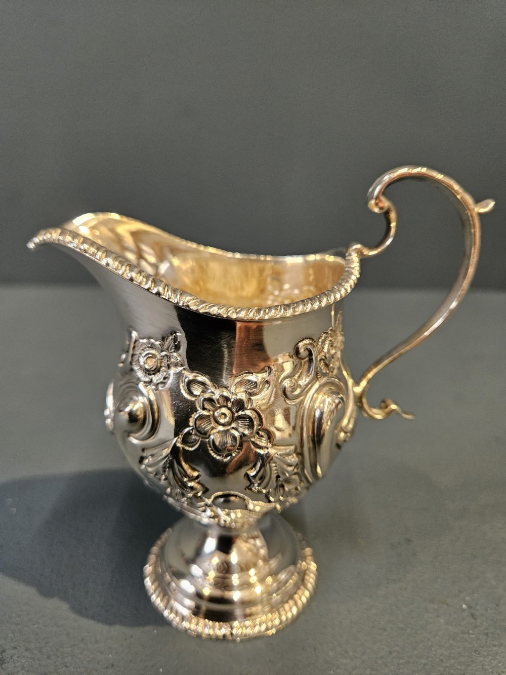 An Edwardian silver cream jug with floral embossing, a high-arched flying scroll handle, and a circular pedestal base.