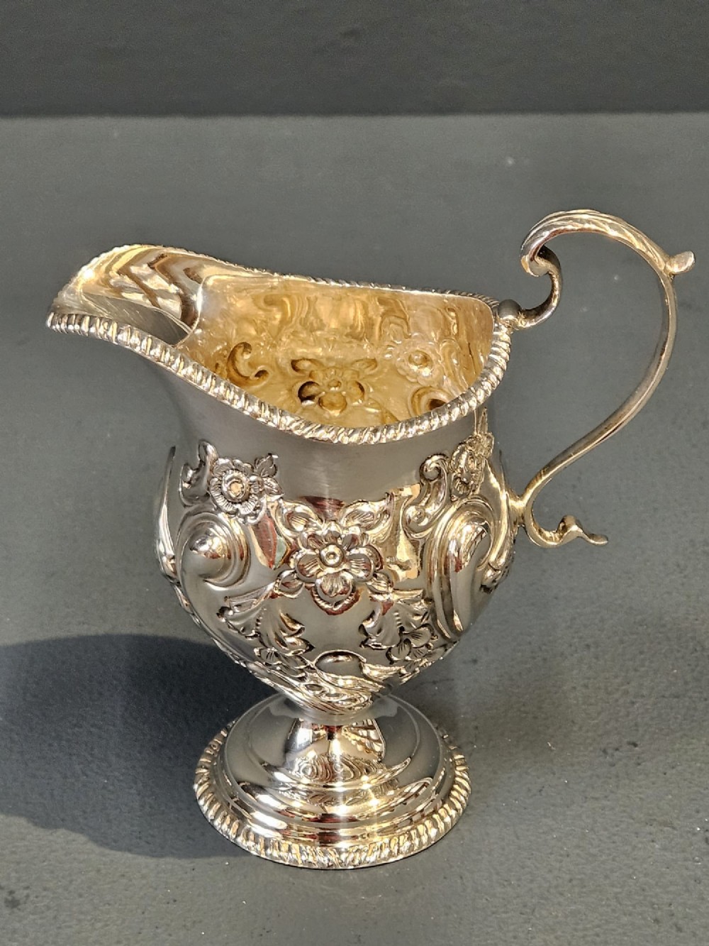 An Edwardian silver cream jug with floral embossing, a high-arched flying scroll handle, and a circular pedestal base. View II