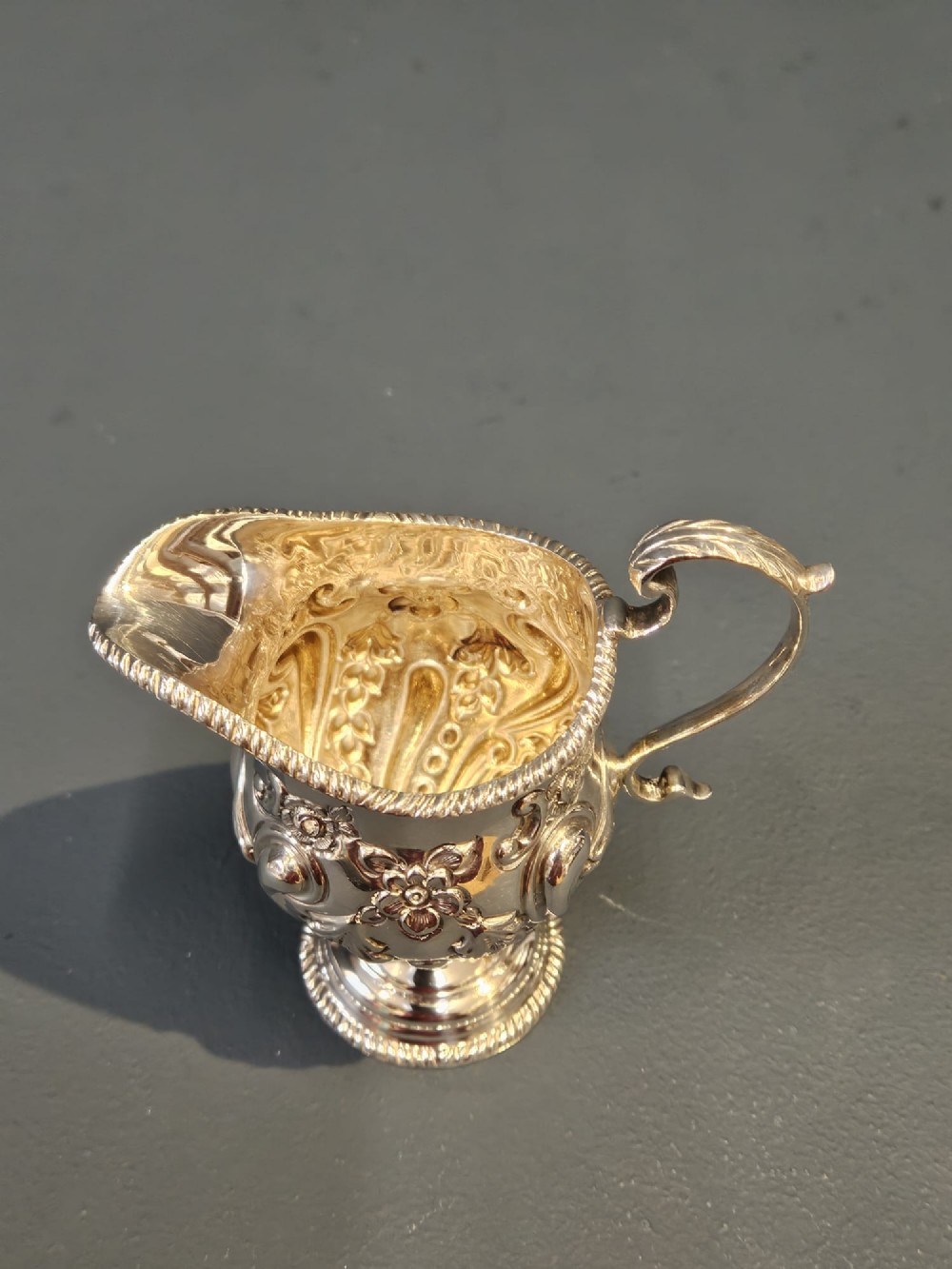 An Edwardian silver cream jug with floral embossing, a high-arched flying scroll handle, and a circular pedestal base. View III