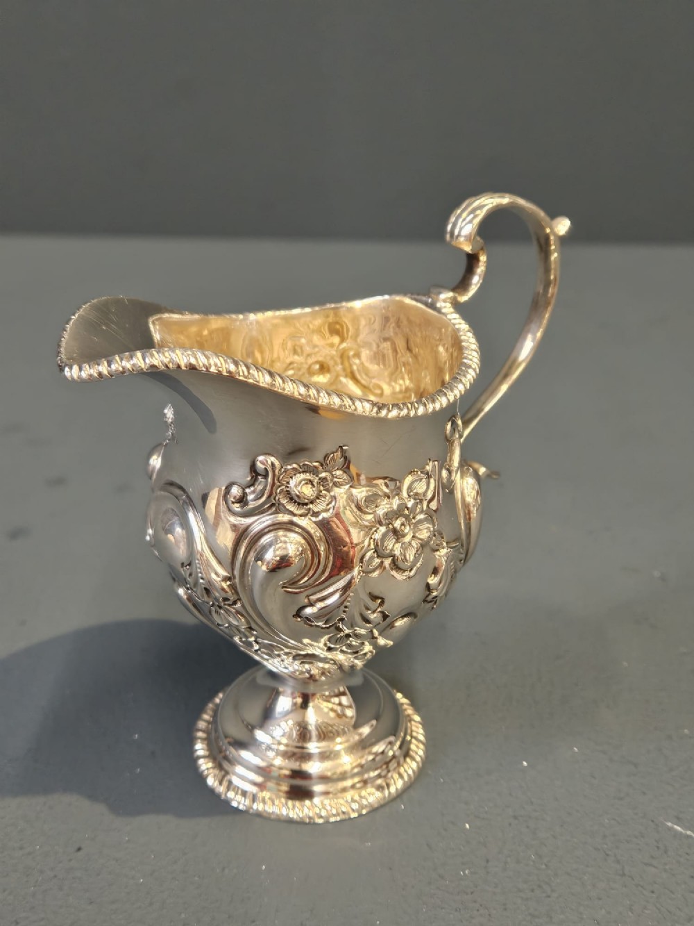 An Edwardian silver cream jug with floral embossing, a high-arched flying scroll handle, and a circular pedestal base. View IV