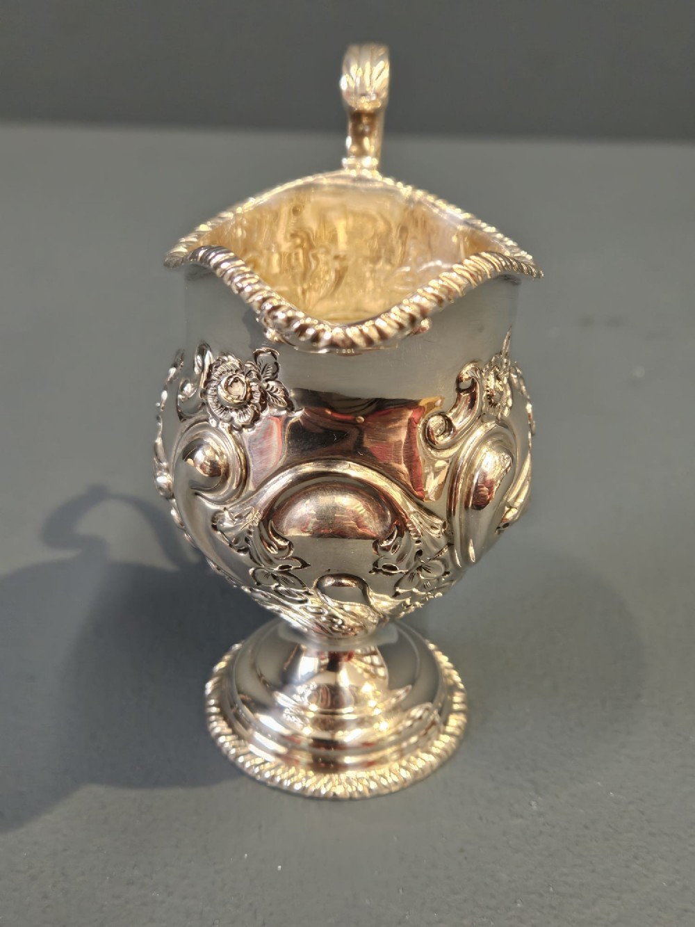 An Edwardian silver cream jug with floral embossing, a high-arched flying scroll handle, and a circular pedestal base. View V