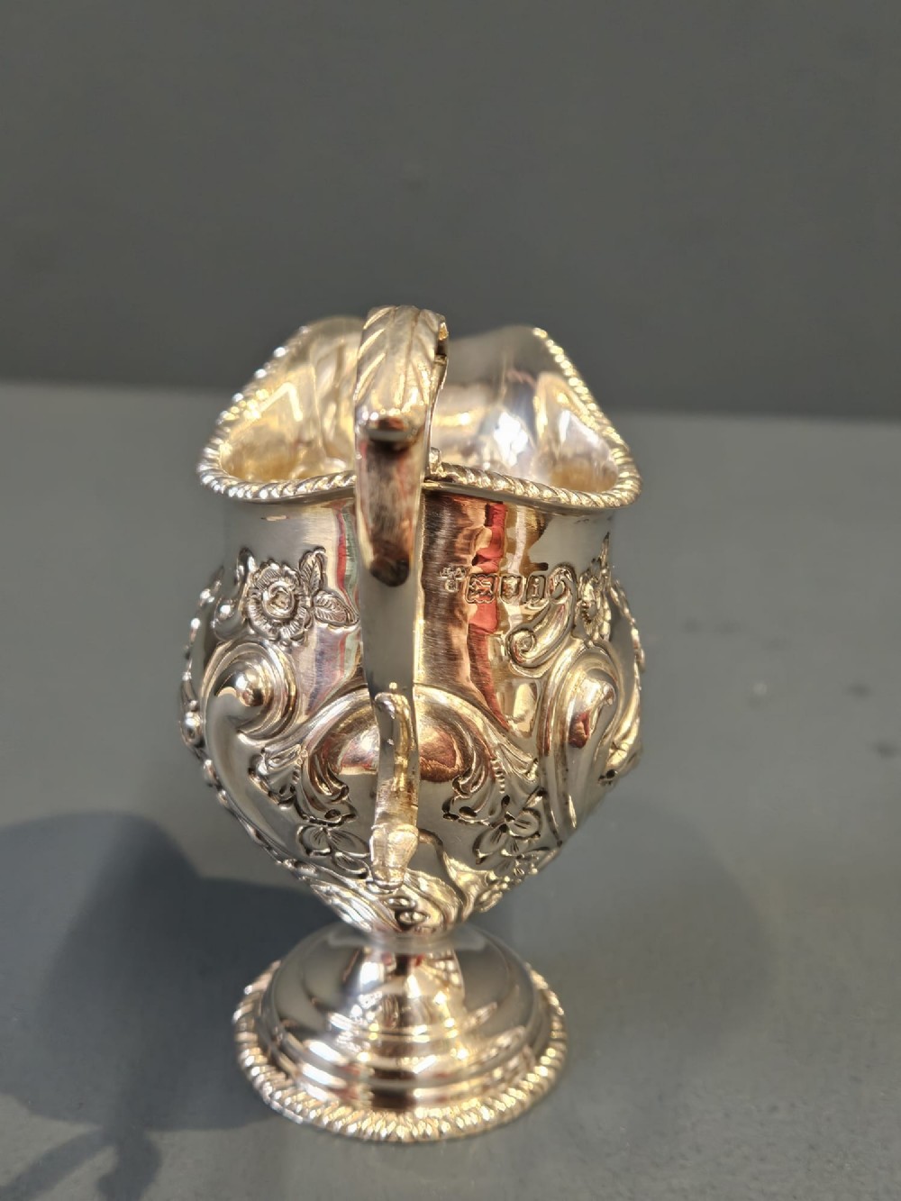An Edwardian silver cream jug with floral embossing, a high-arched flying scroll handle, and a circular pedestal base. View VI