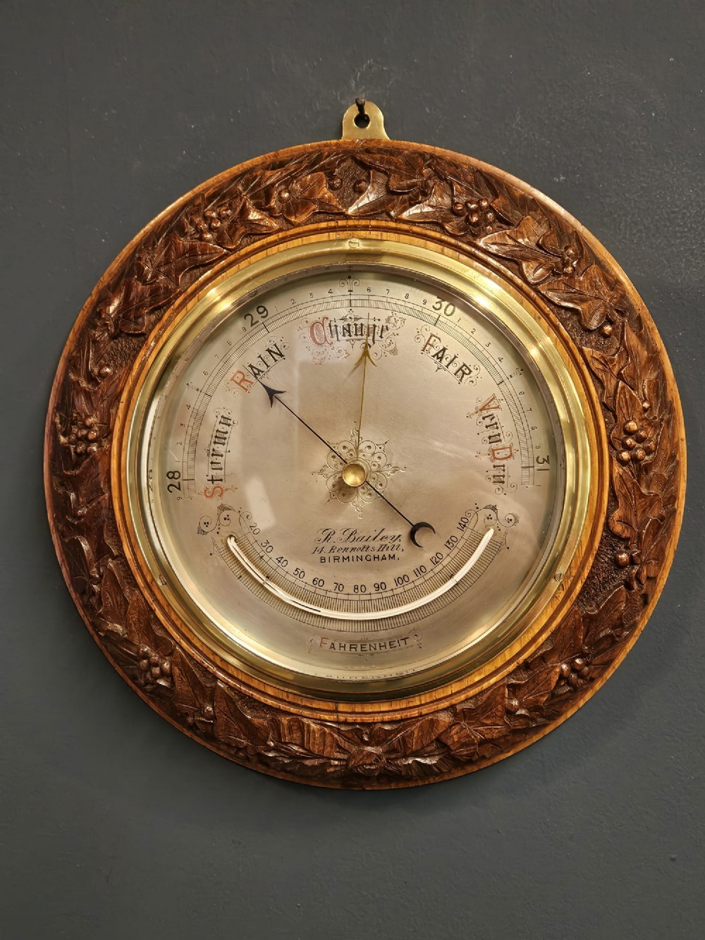 A circular Victorian aneroid barometer featuring a hand-carved oak surround with an ivy wreath pattern and a brass-rimmed silvered dial.