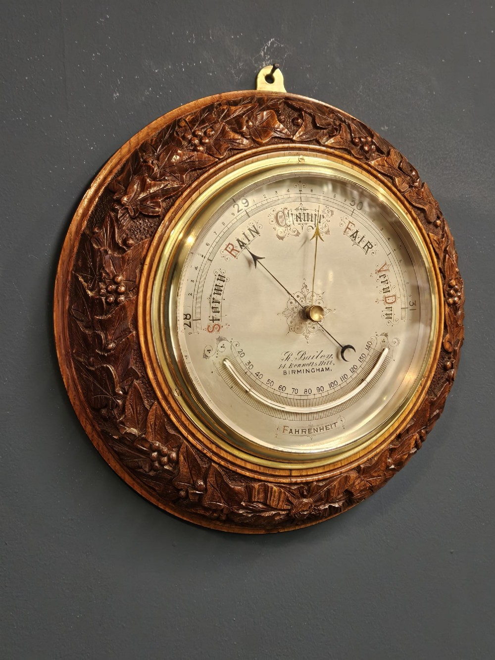 A circular Victorian aneroid barometer featuring a hand-carved oak surround with an ivy wreath pattern and a brass-rimmed silvered dial. View II