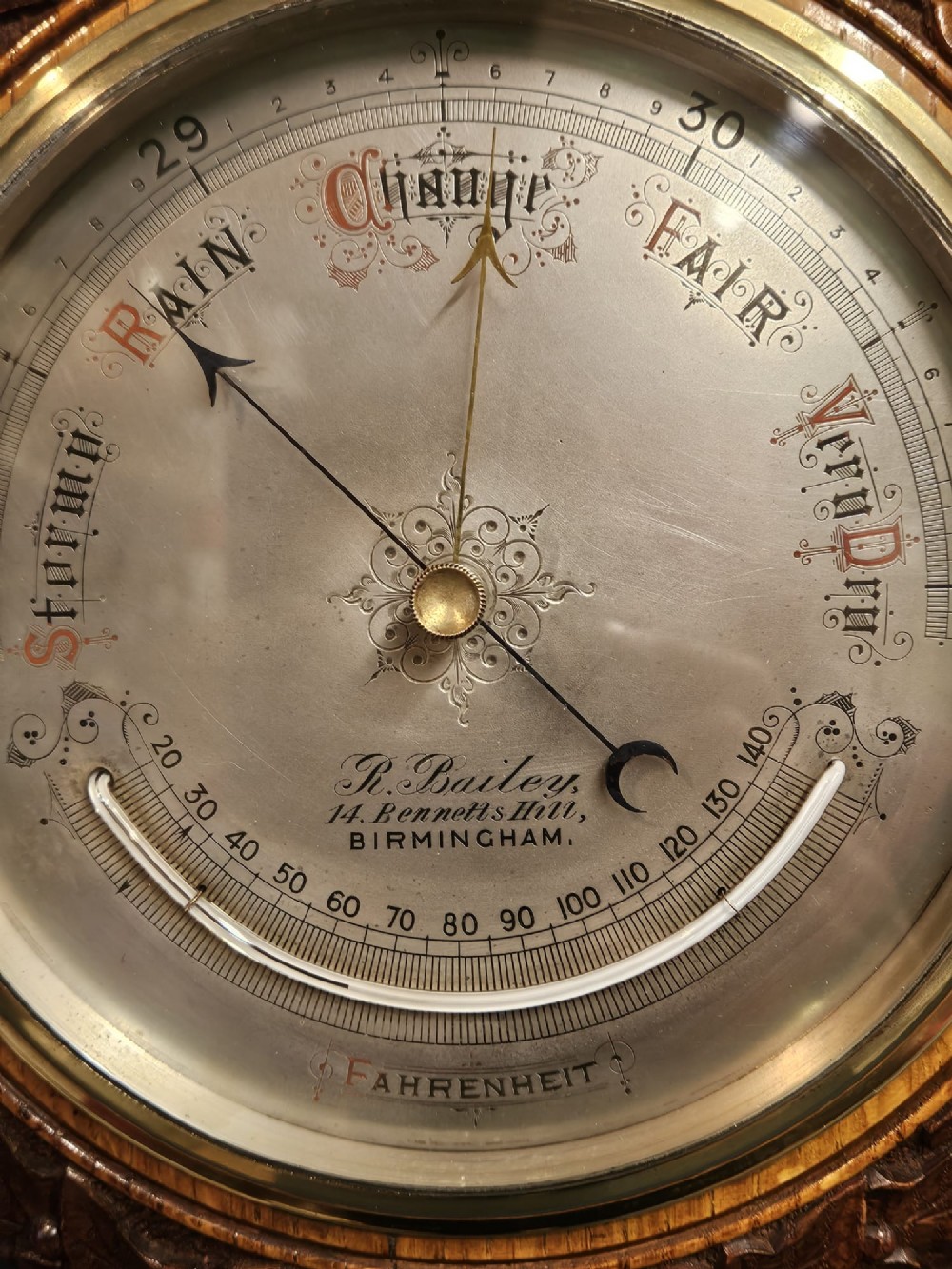 A circular Victorian aneroid barometer featuring a hand-carved oak surround with an ivy wreath pattern and a brass-rimmed silvered dial. View III