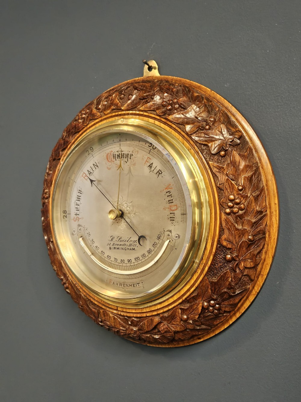 A circular Victorian aneroid barometer featuring a hand-carved oak surround with an ivy wreath pattern and a brass-rimmed silvered dial. View IV