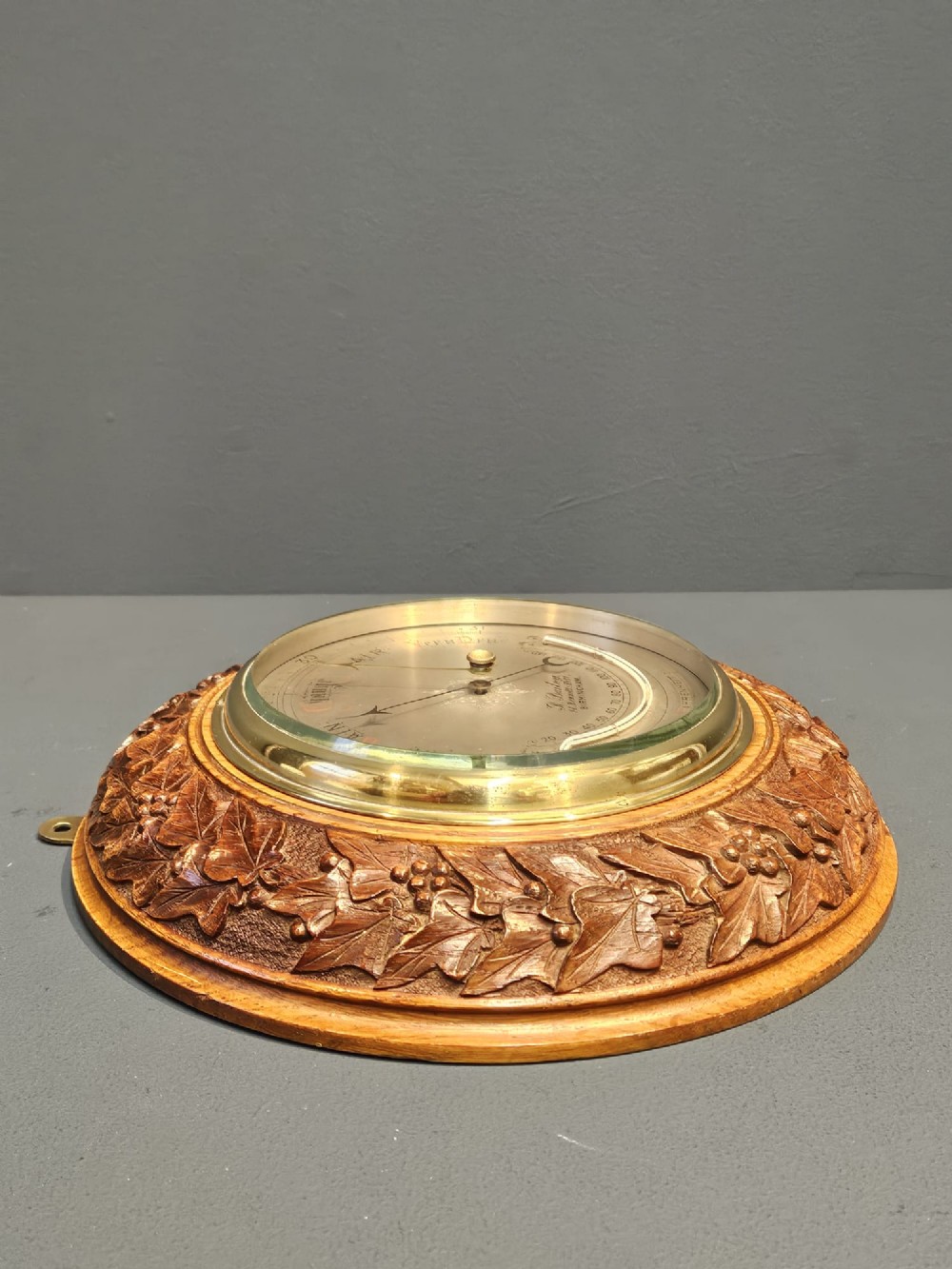 A circular Victorian aneroid barometer featuring a hand-carved oak surround with an ivy wreath pattern and a brass-rimmed silvered dial. View V
