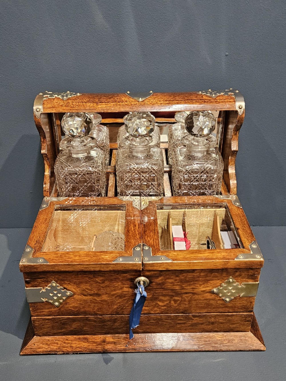 A Victorian oak tantalus featuring brass corner mounts, three cut-glass decanters with faceted stoppers set against a mirrored back, and forward-folding compartments for gaming accessories.