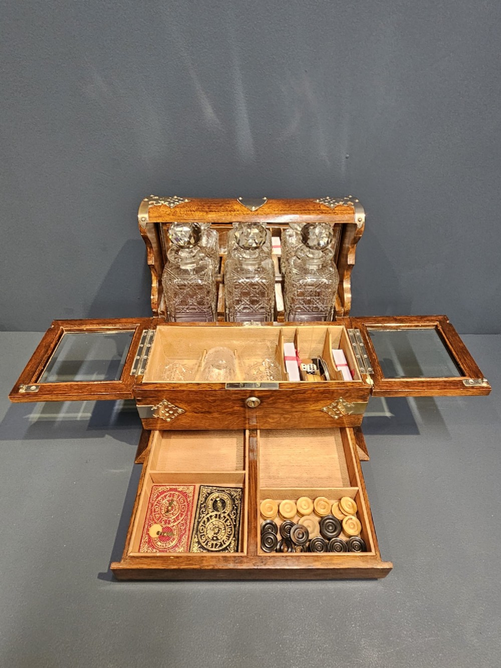 A Victorian oak tantalus featuring brass corner mounts, three cut-glass decanters with faceted stoppers set against a mirrored back, and forward-folding compartments for gaming accessories. View II